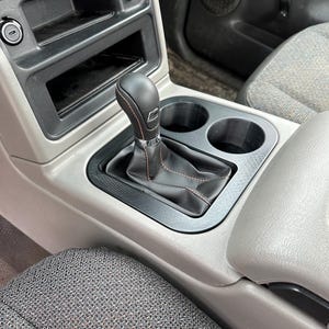 May include: A black leather gear shift knob with red stitching in a gray car interior. The gear shift is surrounded by a black plastic console with two cup holders.