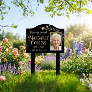 Puede incluir: Una estaca de jardín conmemorativa negra con el texto "Forever Loved" y "Margaret Collins 1950-2023 A Life Full of Love". Presenta una foto enmarcada de una mujer y está colocada en un jardín con flores.