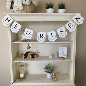 May include: A white wooden hutch with a banner that reads "He is Risen" in black letters with green foliage. The banner is strung across the top shelf of the hutch. The hutch has two shelves and two drawers. There are decorative items on the shelves, including a wooden nativity scene, a cross, and a small potted plant.