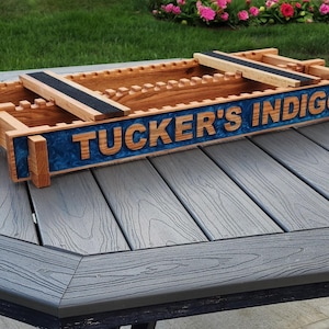 May include: Wooden storage box with the text "TUCKER'S INDIGO" in raised letters. The box has a blue resin inlay and two wooden crossbars with black strips. The box is made of light brown wood and sits on a gray table.
