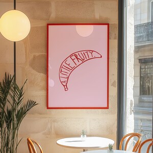 Puede incluir: Una impresión artística enmarcada con un fondo rosa, una ilustración de plátano rojo y el texto "A Little Fruity". La obra de arte está en un marco rojo y cuelga de una pared texturizada.