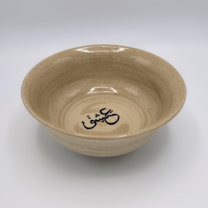 May include: A beige ceramic bowl with a wide rim and a slightly irregular shape. The interior features a spiral pattern and a black script design. The bowl is suitable for serving food or as a decorative piece.