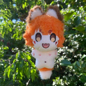 Puede incluir: Un llavero de peluche con un personaje estilizado de pelaje naranja brillante, orejas puntiagudas con puntas marrones y cara blanca. El personaje tiene ojos marrones bordados, una boca sonriente con un colmillo visible y cuerpo blanco.