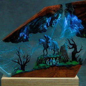Può includere: Un'opera d'arte in resina unica con una scena fantasy con un cavaliere a cavallo, alberi e figure. L'opera d'arte è racchiusa in resina trasparente con una base e una cornice in legno, che mostra effetti vorticosi blu e bianchi.