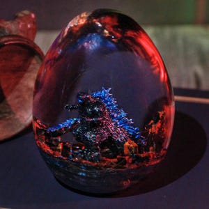 May include: An egg-shaped resin sculpture with a miniature Godzilla figure inside. The sculpture is primarily blue, red, and black, with a detailed cityscape at the base. The sculpture is displayed on a dark blue surface.