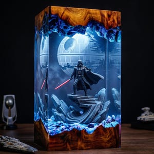 May include: A rectangular resin and wood lamp featuring a Darth Vader figurine standing on a rocky base with a glowing red lightsaber. The interior displays a blue-lit Death Star against a dark background, encased in clear resin with natural wood edges.