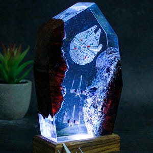 Peut inclure: Sculpture en résine lumineuse représentant le Faucon Millenium et des chasseurs X-wing dans une scène spatiale. La sculpture est posée sur une base en bois. Un petit modèle de chasseur spatial est devant.