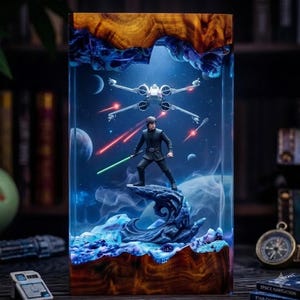 May include: A rectangular resin lamp featuring a wooden frame at the top and bottom. Inside the clear resin, a miniature figure of a Jedi holding a green lightsaber stands on a rock, with an X-wing starfighter flying above in a blue space scene.