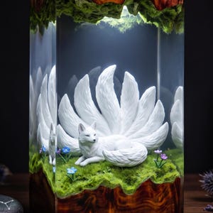 May include: A clear resin block containing a white nine-tailed fox figurine resting on a patch of green moss. Small blue and purple flowers are embedded in the moss. The base of the block is made of polished dark brown wood with a natural grain.