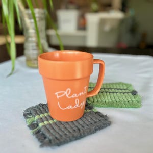 May include: Orange ceramic mug with a white handle and the text "Plant Lady" on the front. The mug is sitting on a gray and green woven coaster.