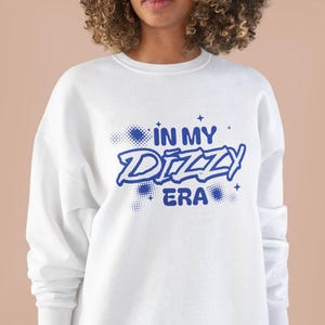 Puede incluir: Sudadera blanca con texto azul que dice "In My Dizzy Era" en una fuente de estilo graffiti.