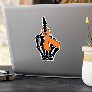 May include: A black skeleton hand giving the middle finger with an orange ribbon tied around the index finger.