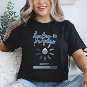 May include: Black t-shirt with the text "healing in progress" in a light blue script. The shirt features a graphic of a brain with a loading bar, suggesting a theme of mental health and self-improvement. The shirt is worn by a person.