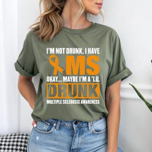 May include: An olive green t-shirt featuring white and orange text that reads: I'M NOT DRUNK, I HAVE MS OKAY... MAYBE I'M A 'LIL DRUNK MULTIPLE SCLEROSIS AWARENESS. The design includes an orange awareness ribbon graphic next to the letters MS.