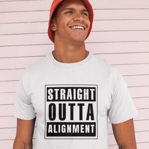 May include: A white short-sleeved t-shirt featuring a black rectangular graphic with the text STRAIGHT OUTTA ALIGNMENT in bold capital letters, paired with a solid orange bucket hat. The design is centered on the chest of the garment.