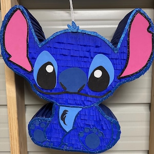 May include: Blue and pink Stitch character pinata. The pinata is made of tissue paper and has a blue body, pink ears, and black eyes.