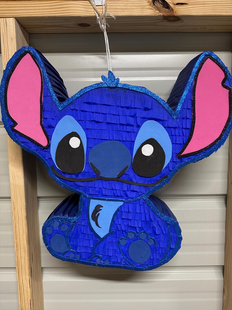 May include: Blue and pink Stitch character pinata. The pinata is made of tissue paper and has a blue body, pink ears, and black eyes.