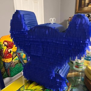 May include: A blue pinata shaped like a cartoon character. The pinata is made of tissue paper and has a fringed design. The pinata is sitting on a table with a yellow tablecloth.