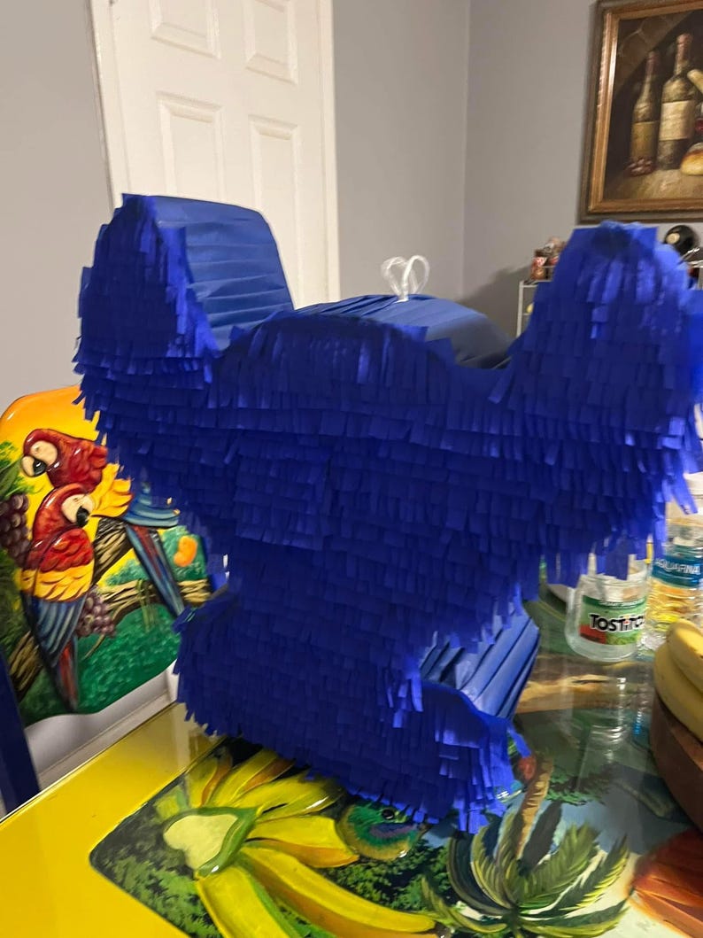 May include: A blue pinata shaped like a cartoon character. The pinata is made of tissue paper and has a fringed design. The pinata is sitting on a table with a yellow tablecloth.