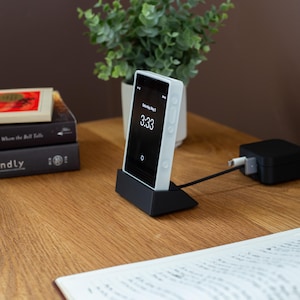 May include: A white smartphone displaying 3:33 on the screen, resting in a black charging dock. The phone is connected to a black power adapter via a black cable. A small green plant in a white pot sits in the background.