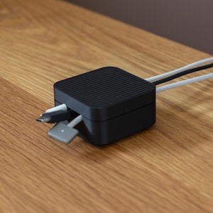 May include: A black square cable organizer with a ridged top. Several cables, including white and gray ones, emerge from the sides. The organizer sits on a wooden surface.