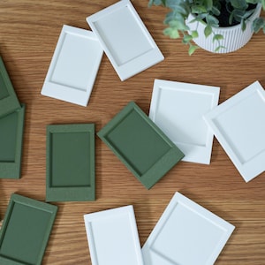 May include: A collection of rectangular photo frame magnets in solid white and matte olive green. Each frame features a raised border and a recessed center, designed to hold small photographs or notes securely on a metallic surface.