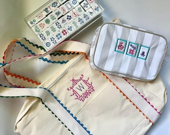 May include: A white canvas tote bag with colorful trim and a monogrammed "W" in a pink floral design. The bag is next to a gray and white striped pouch with embroidered Chinese characters. A clear plastic box filled with colorful mahjong tiles is also in the image.