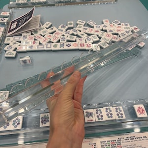 May include: A clear acrylic Mahjong tile rack with a set of white tiles featuring various symbols and designs. The tiles are arranged on a light blue surface, with some tiles in the foreground and others scattered in the background. A printed sheet is also visible.