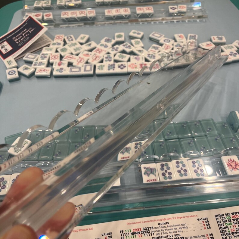 May include: Clear acrylic Mahjong tile racks with curved supports are featured in this image. The racks are positioned above a collection of Mahjong tiles, some with floral and geometric designs. The game set includes tiles with the word "Joker" on them.