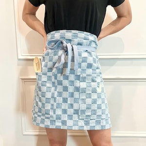 Puede incluir: Un delantal a cuadros azules y blancos con una cinta azul claro y dos bolsillos. Una cuchara de madera está en el bolsillo izquierdo.