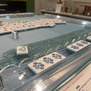 May include: A close-up of a Mahjong game in progress. The tiles are white with blue and green markings. The game is set on a light blue surface with clear acrylic dividers. Some tiles are arranged in rows, while others are scattered.