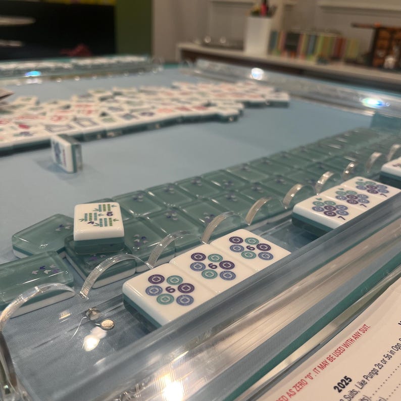 May include: A close-up of a Mahjong game in progress. The tiles are white with blue and green markings. The game is set on a light blue surface with clear acrylic dividers. Some tiles are arranged in rows, while others are scattered.