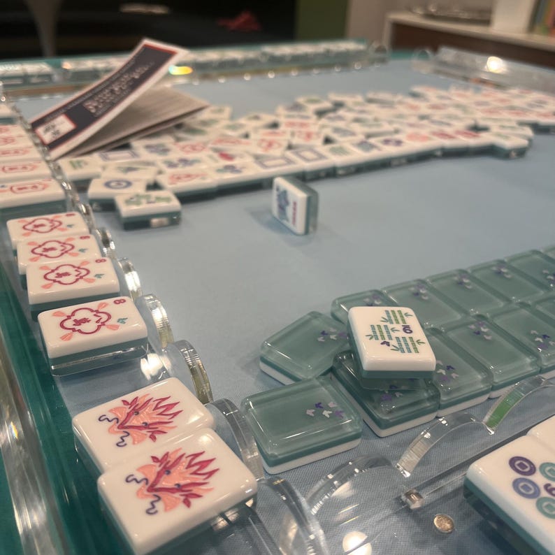 May include: A Mahjong set with white tiles featuring various symbols and characters. The tiles are arranged on a light blue surface, with some tiles stacked and others scattered. The set includes clear, green-tinted tile holders.