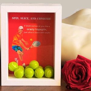 Op de afbeelding: Een wit fotolijstje met een rode achtergrond en een illustratie van een tennisspeler. De tekst in het lijstje luidt "SPIN, SLICE, AND CONQUER! If 2025 comes at you like a crazy topspin, just slice it back with style!" Er zijn verschillende gele tennisballen in het lijstje.