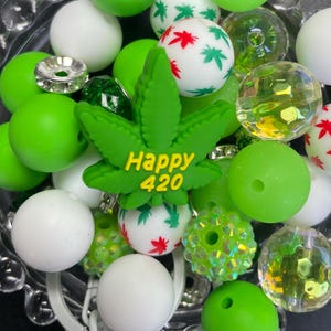 May include: A collection of green and white beads, including round and faceted shapes. A green cannabis leaf with the text "Happy 420" is the focal point. Some beads have cannabis leaf patterns in red and green.