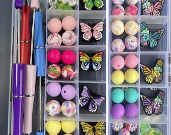 May include: A clear plastic organizer box containing various craft supplies, including three metal pens with colorful barrels, assorted round silicone and patterned beads, and multiple butterfly-shaped silicone charms in vibrant colors.