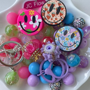 Op de afbeelding: Een verzameling kleurrijke knutselkralen en accessoires, waaronder drie ronde smiley-bedels met bloemen- en aardbeienpatronen, een roze roosvormige bedel en een paarse sleutelhangerring, alles op een witte, geschulpte schaal.
