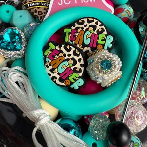 Puede incluir: Un anillo de silicona turquesa con dos dijes de "Teacher" de estampado de leopardo, un dije de pedrer&iacute;a turquesa y una variedad de cuentas de colores, incluyendo turquesa, rosa, blanco y negro.