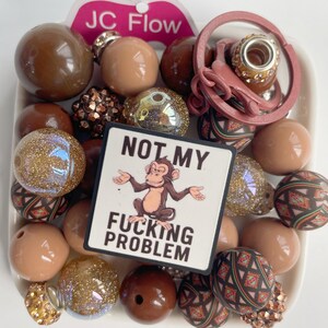 May include: A collection of beads in various shades of brown, tan, and gold, including some with glitter and patterned designs. A small sign with a monkey graphic and the text "NOT MY FUCKING PROBLEM" is also present.