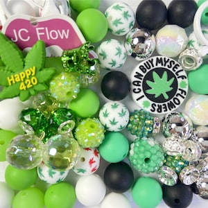 Pode incluir: Uma coleção de contas e berloques verdes, brancos e pretos. Algumas contas apresentam padrões de folhas de cannabis. Um berloque diz "I CAN BUY MYSELF FLOWERS" e outro diz "Happy 420".