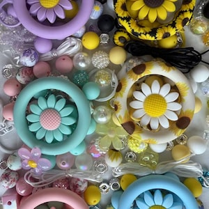 May include: A collection of colorful silicone teething toys with daisy and sunflower designs. The toys are surrounded by a variety of beads, including pearl, wooden, and plastic beads.