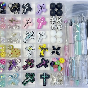 May include: A clear plastic organizer filled with various craft supplies. The compartments contain beads, crosses with the word "Jesus", and pens in shades of pink, green, and black. The beads are in various colors and styles.