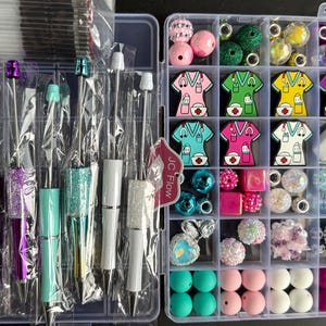 May include: Two clear plastic storage containers filled with colorful beads, charms, and pens. The container on the left has a variety of glitter pens with pink, purple, and blue accents. The container on the right has a variety of colorful beads, charms, and a white retractable badge holder.