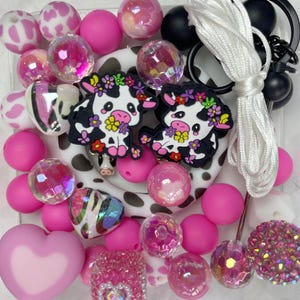 May include: A collection of pink and white beads, including round, heart-shaped, and faceted designs. Two cow-shaped charms with floral crowns are also present, along with a white cord and black hardware. The beads vary in size and texture.