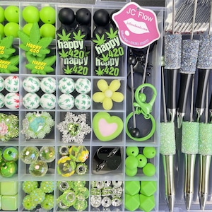 May include: A clear plastic storage box filled with various green, black, and white beads, charms, and other jewelry making supplies. The box also includes four black and green pens with glitter accents. The text "JC Flow" and "jeflowbeads.com" is printed on a pink sticker on the box.