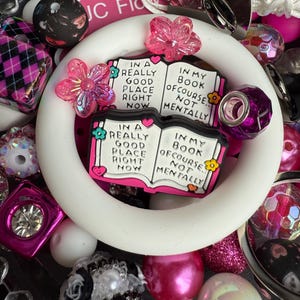Puede incluir: Una pulsera de silicona blanca con un dije de libro rosa y blanco que dice "In a really good place right now. In my book of course, not mentally." La pulsera est&aacute; rodeada de varias cuentas rosas, blancas y negras.