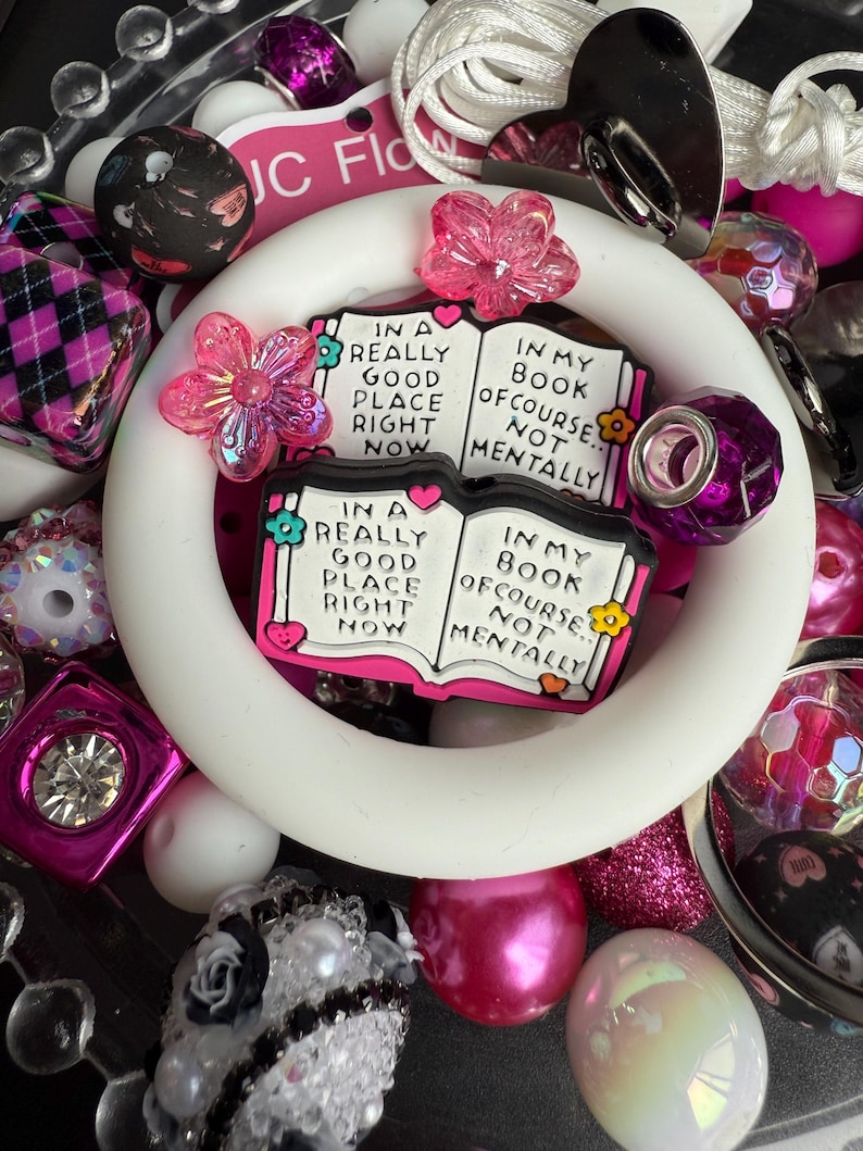 Puede incluir: Una pulsera de silicona blanca con un dije de libro rosa y blanco que dice "In a really good place right now. In my book of course, not mentally." La pulsera est&aacute; rodeada de varias cuentas rosas, blancas y negras.
