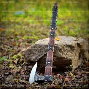 May include: A handcrafted axe with a wooden handle and a silver blade. The handle is wrapped in black leather with brass accents. The axe is leaning against a large rock, with a natural background of leaves and grass.