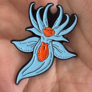 May include: A metal enamel pin shaped like a stylized sea slug or nudibranch. The creature features a light blue body with black outlines and vibrant orange textured accents on its dorsal area and central body, set against a black background.