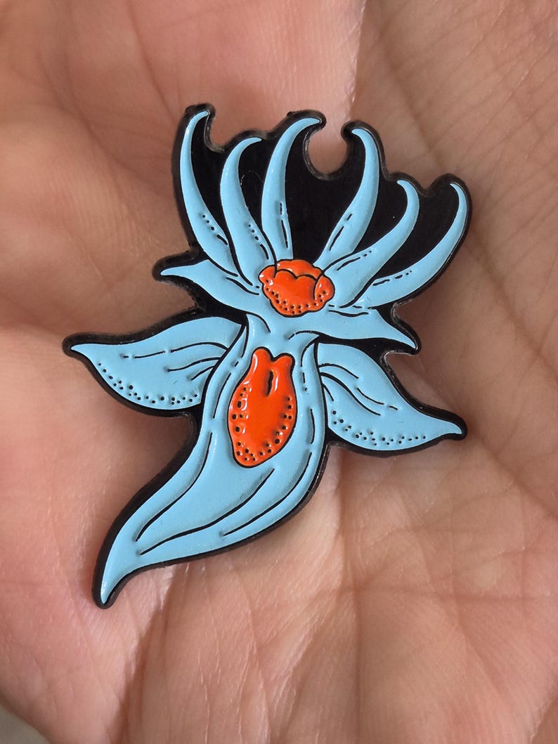 May include: A metal enamel pin shaped like a stylized sea slug or nudibranch. The creature features a light blue body with black outlines and vibrant orange textured accents on its dorsal area and central body, set against a black background.